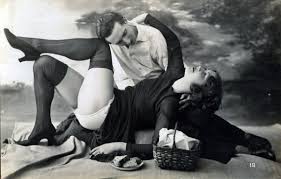 A clothed sitting behind a reclining woman. Her stockings and panties are visible. 